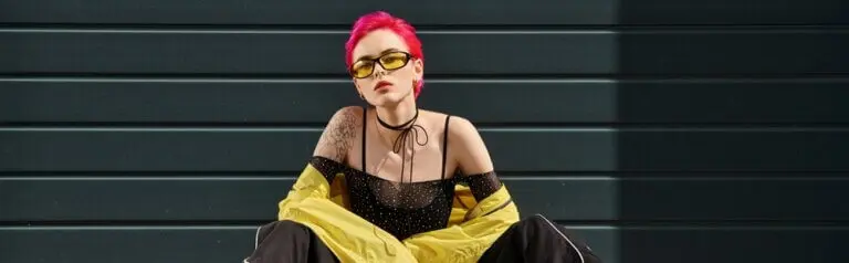 Hip young woman with tattoos piercings and pink hair sitting in front of a black garage door.