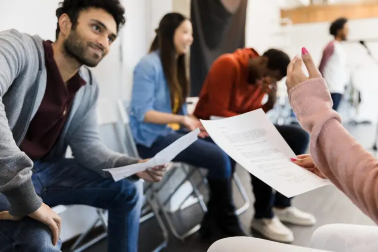 Diverse group of actors at an in-person audition, waiting their turns as one auditions for casting directors. Some look stressed, holding sides.
