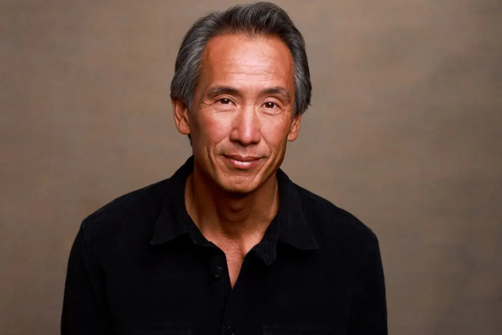 Actor Tinpo Lee standing up and smiling in a black sweater.