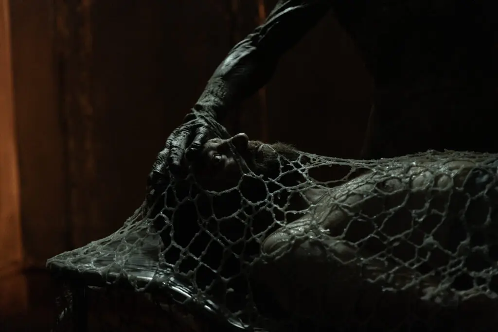 Ross Marquand lying flat in a diark area covered in a coccoon with a n alien hand over his head.