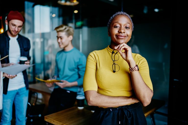 Portrait of african american young woman with eyeglasses in hand smiling at camera while team of students collaborating on background in modern office.Dark skinned female leader holding eyewear