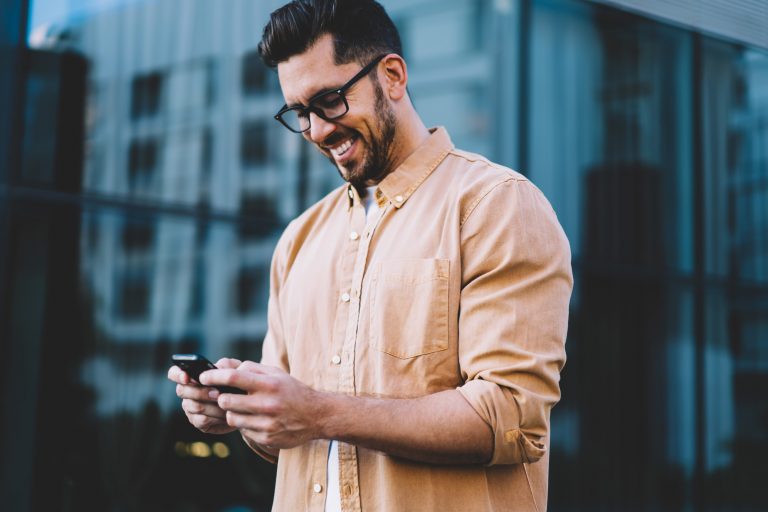 Successful bearded man in eyeglasses for provide eyes protection smiling while reading positive sms from friend connected to 4g,happy male browsing website via smartphone gadget while standing outdoor