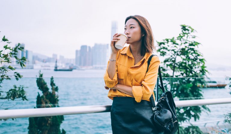 Pensive Japanese hipster student with trendy leather backpack standing at urban setting with caffeine beverage drinking and enjoying coffee taste during Chinese holidays in Hong Kong, tourism