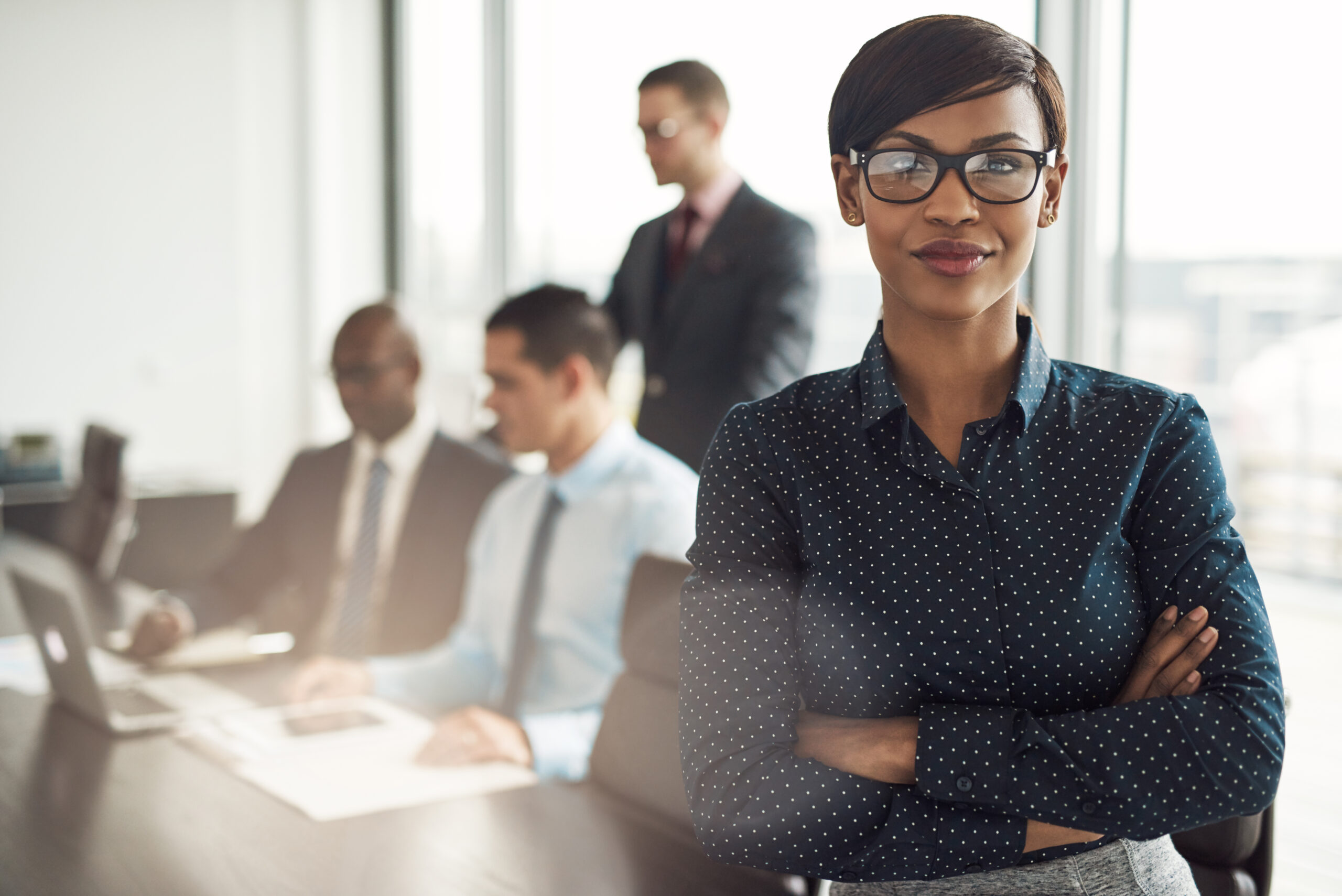 Confident young African businesswoman standing with folded arms smiling at the camera in a boardroom with male colleagues in the background