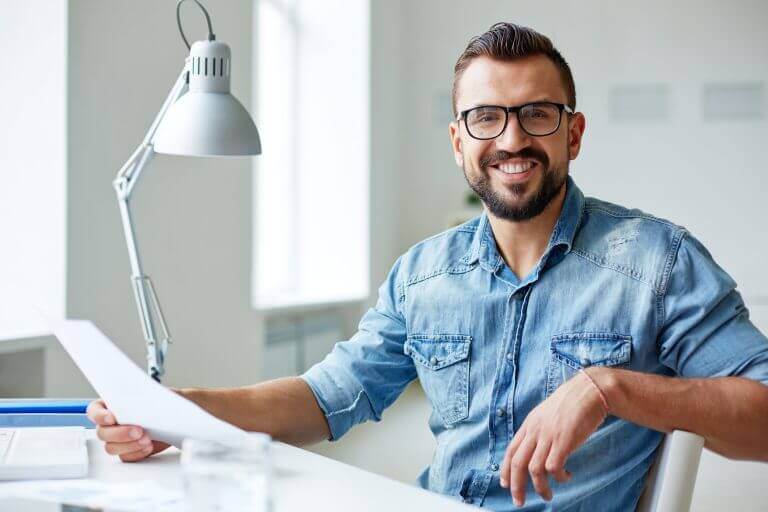 A smiling actor, glasses and blue denim shirt, holding a script at his desk.