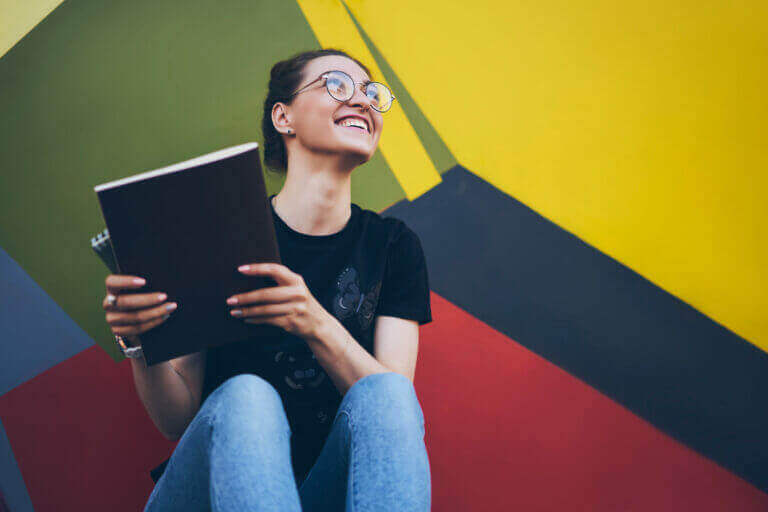 Happy smiling female student in spectacles overjoyed about successfully completed course work in textbook while sitting near colorful wall, cheerful woman feeling excited from information in notepad
