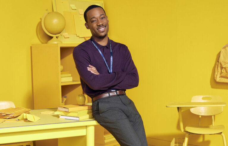 Actor Tyler James Williams leaning against a desk in a yellow classroom.