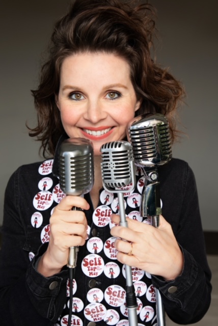 Audrey Moore smiling and holding three microphones while wearing a black shirt adorned with lots of Self Tape May buttons on a black shirt.