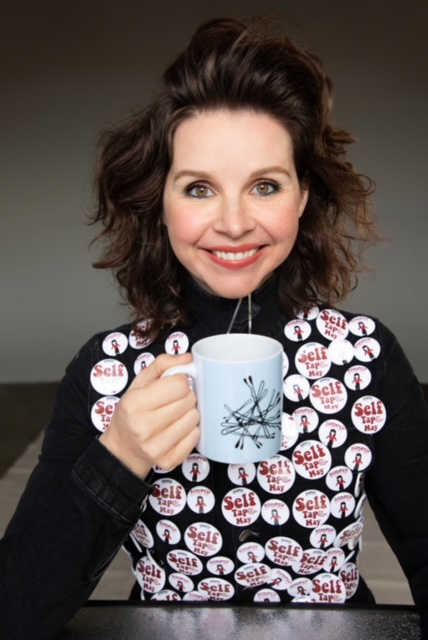 Audrey Moore smiling and drinking coffee with lots of self tape may buttons on a black shirt.