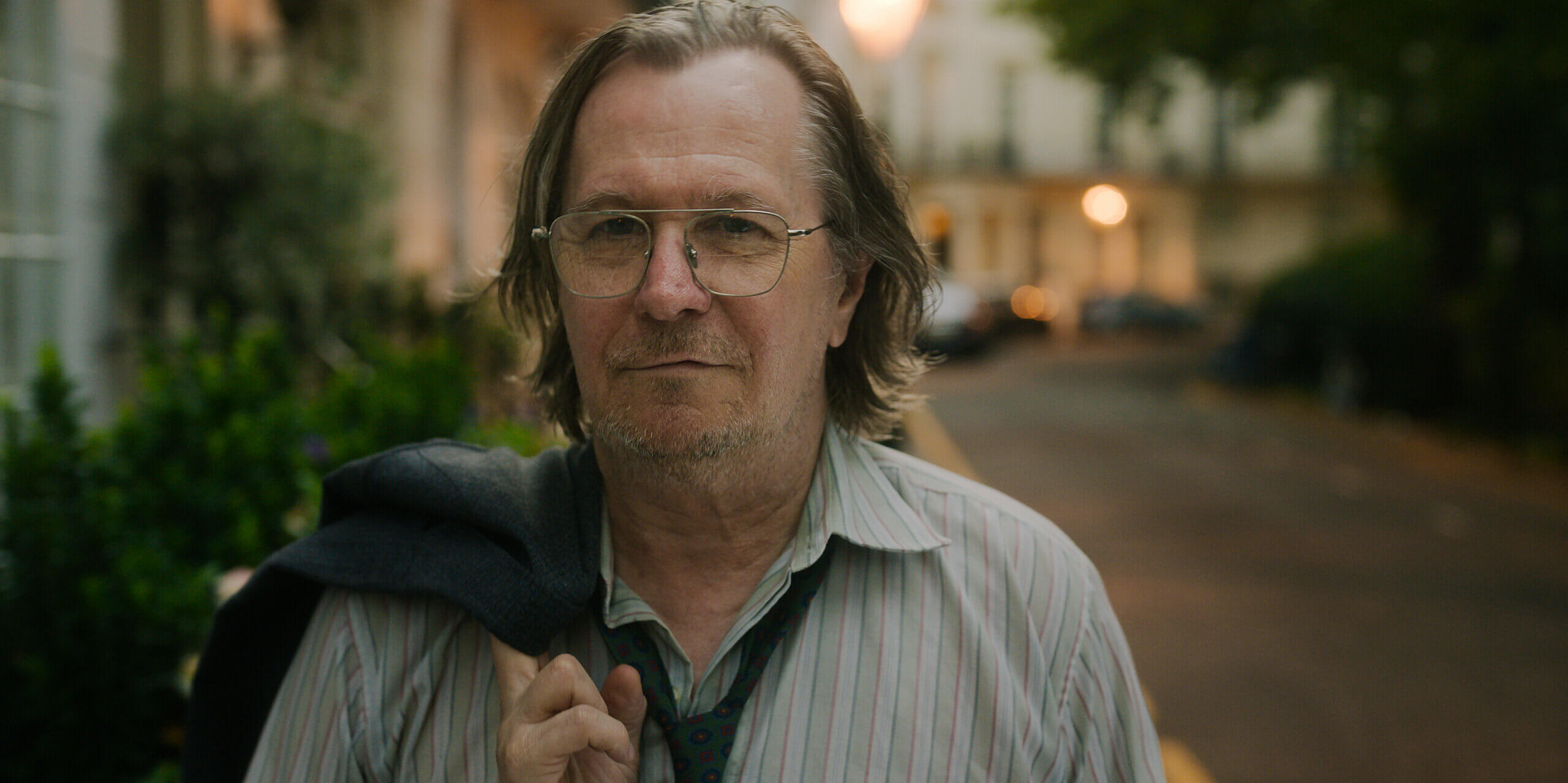 Actor Gary Oldman walking outside in a gray shirt and black tie.