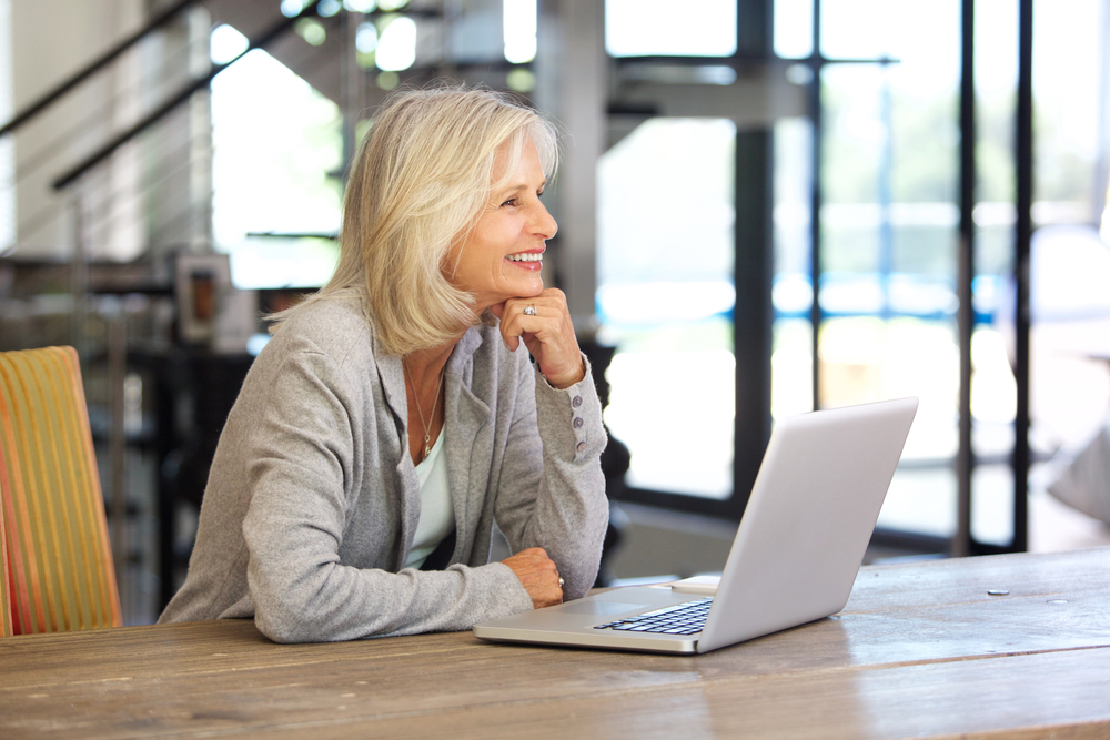 Older woman in a grey cardigan on her laptop smiling.