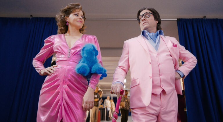 Elizabeth Banks and Zach Galifianakis in matching pink outfits holding a blue teddy bear.