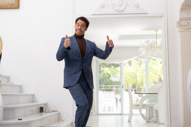 Ike Barinholtz in a suit giving a thumbs up in a nice house.