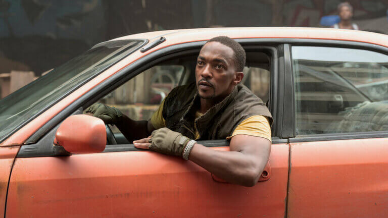 Actor Anthony Mackie driving an orange car outside.