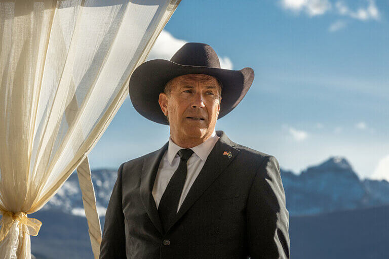 Kevin Costner in the show Yellowstone wearing a suit with a cowboy hat outside.