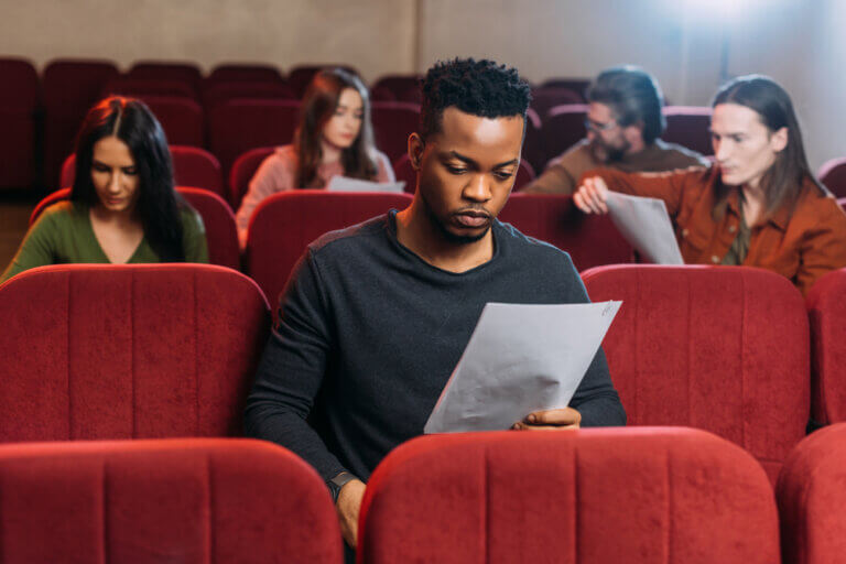 African American actor, surrounded by other actors, reading a script in a theater chair.