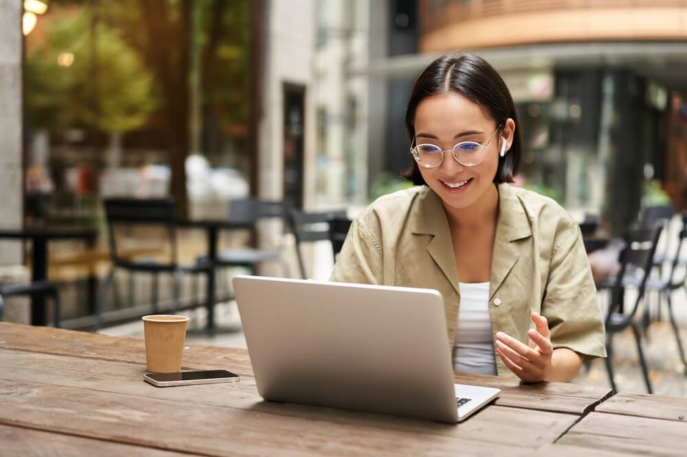 Happy Asian woman working on her laptop outside a bustling city cafe.