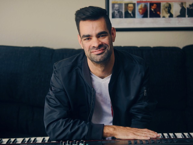 Composer Michael Wilson sitting on the couch in a blue jacket smiling.