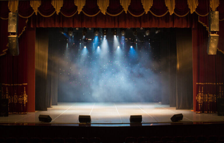 An empty theater stage lit up by spotlights and smoke.