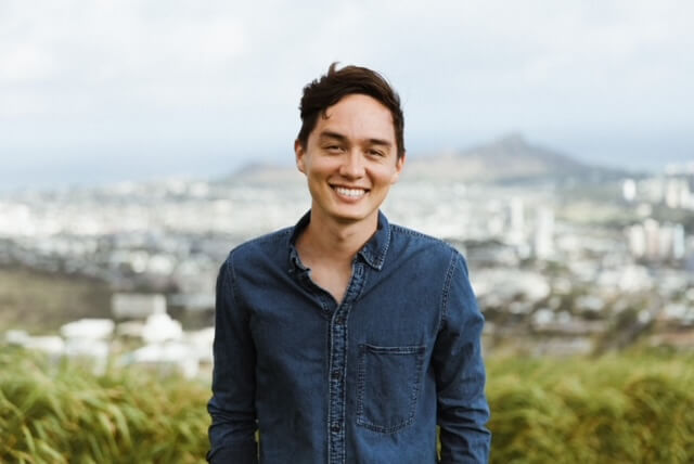 Chris Kekaniokalani Bright standing at the top of a mountain with a beautiful city view behind him.
