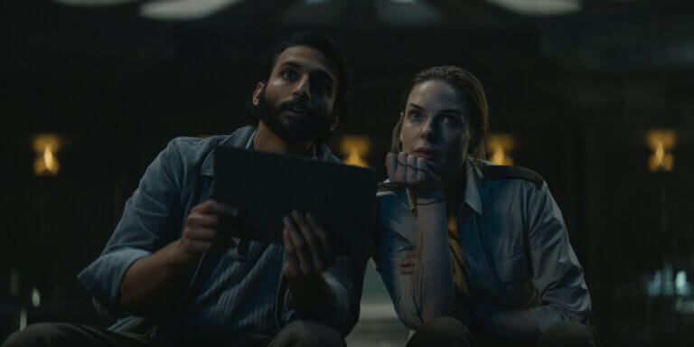 Avi Nash and Rebecca Ferguson holding a notepad staring.
