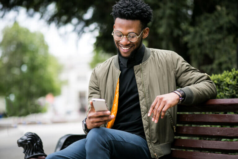 Young African American man sitting on a park bench happily on his phone.
