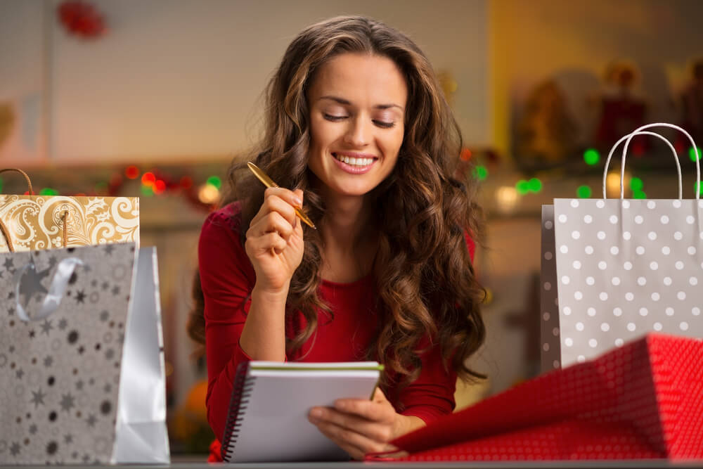 Woman making s holiday savings list, surrounded by bags of gifts.