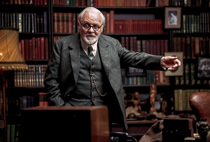 Sir Anthony Hopkins in a suit surrounded by books pointing.