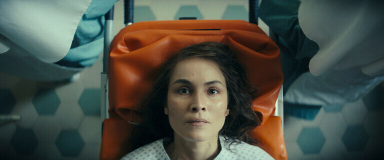 Noomi Rapace on a gurney bed looking blankly at the ceiling.