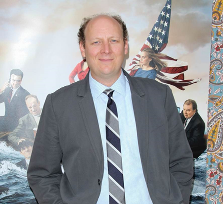 Dan Bakkedahl smiling in a suit behind a war painting.