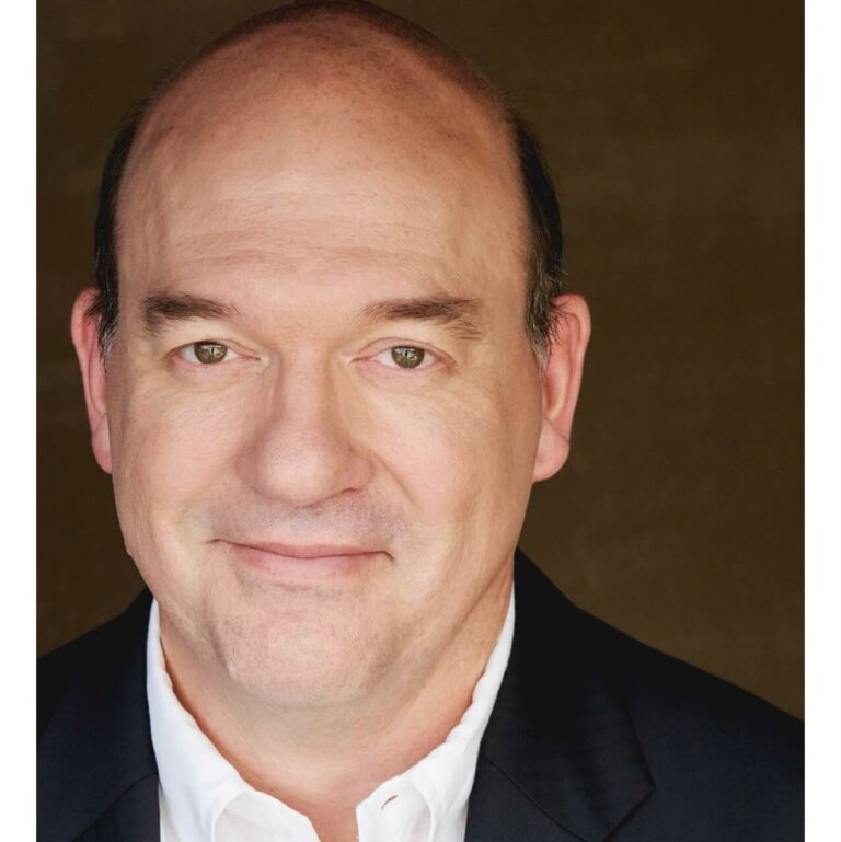 John Carroll Lynch in a button down shirt and jacket smiling.