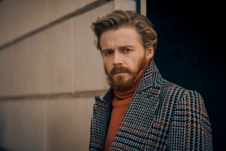 Jack Lowden in a warm checkered jacket and burgundy shirt outside.