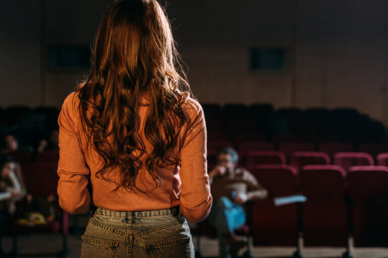 Actress in orange auditioning in front of a casting director in an empty theater.