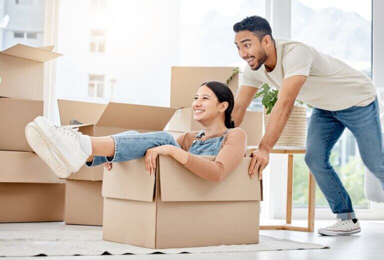 Interracial couple moving. Man pushing a box with his female friend inside laughing.