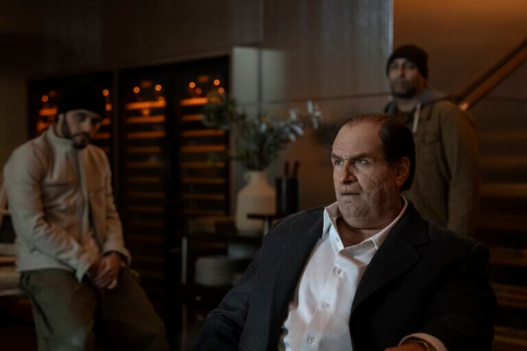 Colin Ferrell as "The Penguin" sitting in a chair with associates inside a hotel.