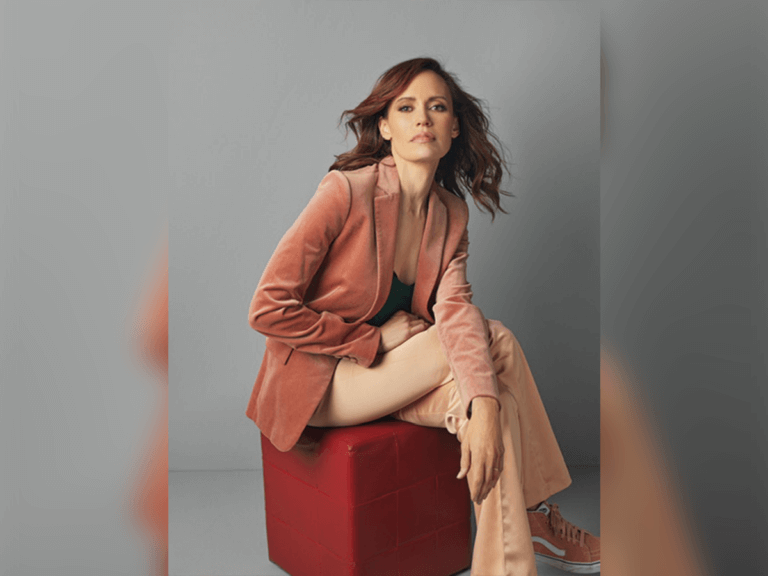 Actress Emily Swallow sitting on a red cube wearing pink outfit.