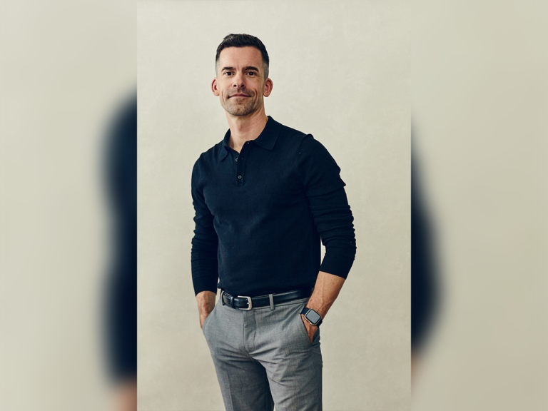 Casting Director Jesse Griffiths standing and smiling in a blue shirt, grey pants inside.