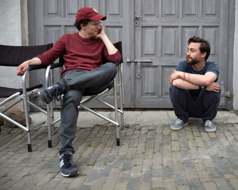 Jesse Eisenberg sitting in a chair and Kieran Culkin squatting on the set of A REAL PAIN.