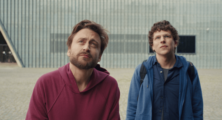Jesse Eisenberg and Kieran Culkin looking up at an industrial warehouse.