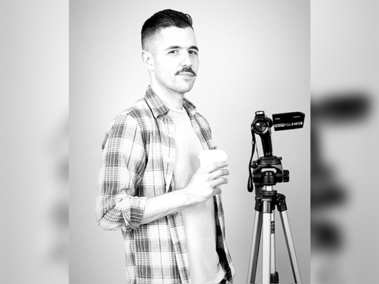 Black and white photo of Matthew Maisto standing next to his camera on a tripod with a cup of coffee.