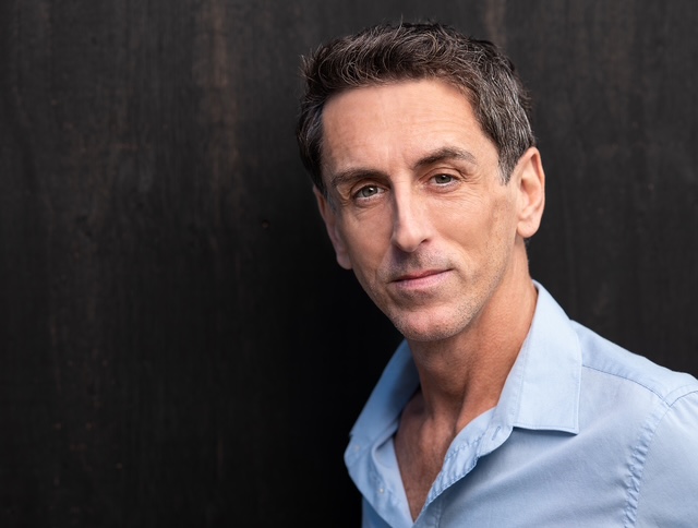 Voice Actor Duff McDonald in a blue shirt smiling inside.