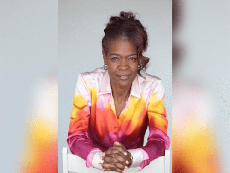 Casting Director Leslie M. Greene smiling in a rainbow blazer at her desk.