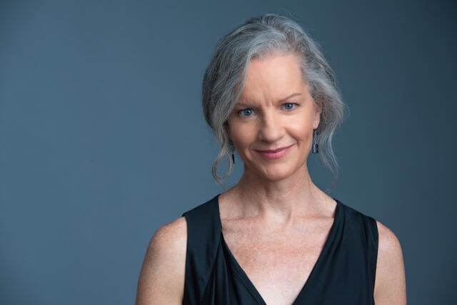 Tracey Hoyt in a black shirt smiling in front of a dark blue background.