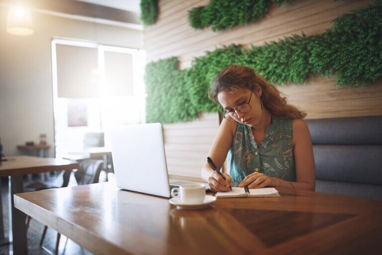Young woman at a coffee shop researching on her laptop and making notes on a pad.