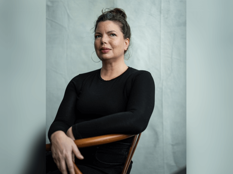 Casting Director Bess Fifer, dressed in black, sitting in a chair in front of a turquoise backdrop.