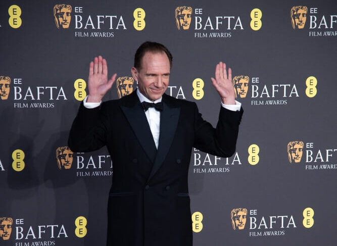 Ralph Fiennes, hands raised, wearing a sharp tuxedo at the BAFTA Awards red carpet event.