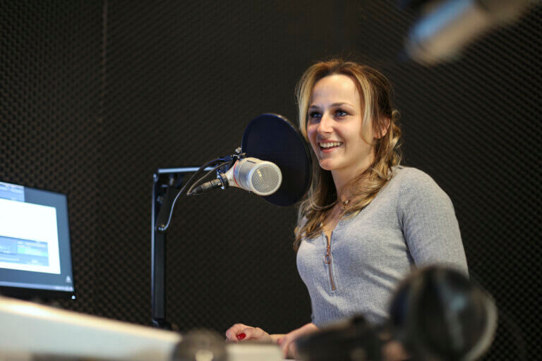 A voice actor in a grey shirt stands at a dekc and reads copy in a large recording studio.