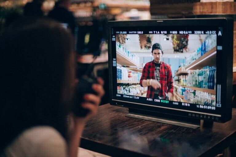 A direct or watches a video monitor on a film set playing back a scene where the lead actor walks through a store in flannel and a backward hat.
