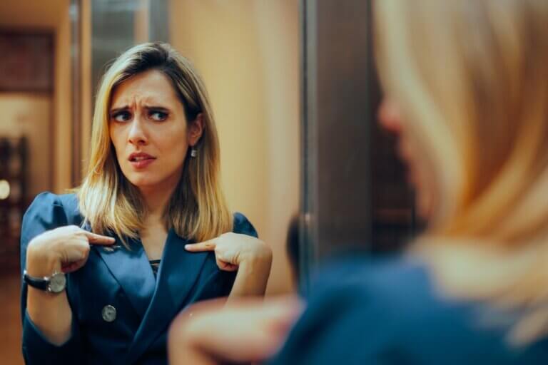 Blonde businesswoman with a blue blazer and watch looking in the mirror going through impostor syndrome.