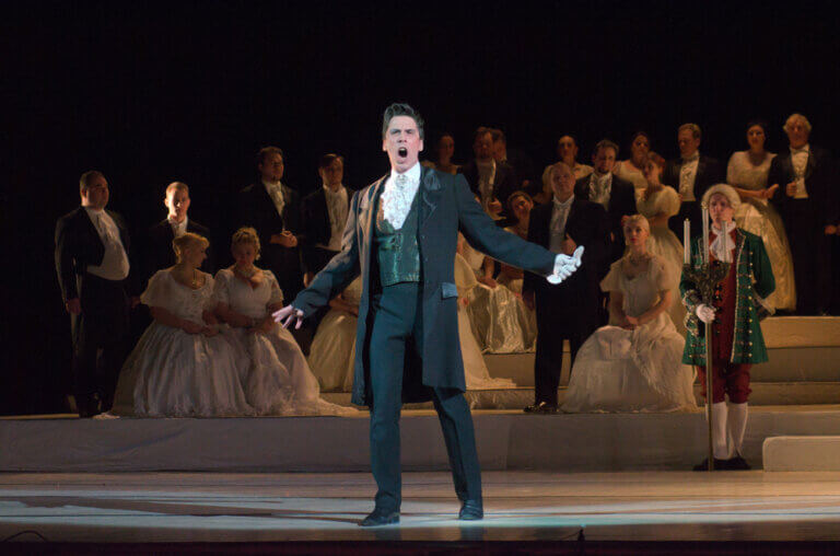 Lead actor singing in a political theater production with an ensemble beihind him in Victorian era costumes.